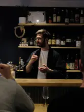 A moody, atmospheric shot taken from a customer's perspective inside an izakaya, featuring a bartender explaining sake. The lighting is intimate and focused on the subject, creating an inviting and educational vibe.