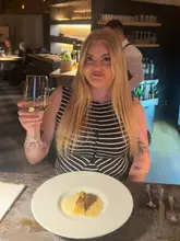 A casual, influencer-style shot taken at a fine dining restaurant. The image features a diner holding a glass of wine with a single course from a tasting menu placed in the foreground.