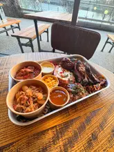 A high-angle shot of a large BBQ platter served on a metal tray, featuring various meats, beans, and sides, set on a wooden table with a view of the waterfront in the background.