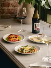 A high-quality, professional food photography shot featuring a spread of small plates and wine at a wine and vinyl bar. The composition is balanced, with a shallow depth of field drawing attention to the foreground dish, while the background elements like the wine bottle and greenery provide context and atmosphere.