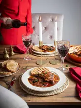 A professionally styled, high-quality food photography shot featuring a rustic stew served with grilled bread and a glass of red wine being poured. The scene is set for an intimate dinner, utilizing warm lighting and elegant props to create an inviting atmosphere.