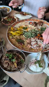 A short, handheld clip showcasing a table spread of Middle Eastern dishes including a large platter with hummus, falafel, and tabbouleh, alongside rice and dips, being enjoyed by a group.