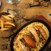 An overhead, high-angle shot of a 'meltdown' dish featuring two burger sliders served in a skillet filled with melted cheese sauce, accompanied by a side of fries and a glass of white wine on a distinctively patterned wooden table.