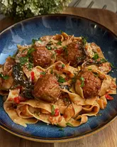 A high-quality, top-down shot of a bowl of creamy Tuscan meatball tagliatelle. The lighting is soft and natural, effectively highlighting the texture of the pasta and the glaze on the meatballs.