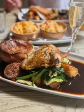 A high-quality, well-lit photograph of a Sunday roast dinner featuring roast chicken, a large Yorkshire pudding, and roast potatoes, served in a pub setting. The image captures the textures of the food effectively, making it look fresh and appetising.