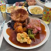 A high-angle shot of a Sunday roast dinner featuring sliced beef, a large Yorkshire pudding, roast potatoes, cauliflower, and red cabbage, served in a pub setting with beer glasses visible.