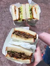 A first-person perspective shot of two sandwiches held in hands, showcasing a chicken schnitzel sando and a steak sando from a street food van.