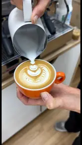 A close-up, high-quality video focusing on the process of pouring latte art into a bright orange coffee cup. The video captures the precision of the pour, resulting in a clean, professional design.
