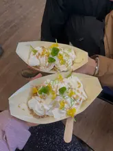 A top-down shot of two people holding small wooden boats containing a dessert garnished with edible flowers and herbs, captured during an outdoor foraging experience.
