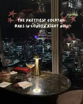 A moody, atmospheric shot of a cocktail bar table at night, featuring a view of the London skyline. The image uses a table lamp to illuminate the drinks and menu, creating a romantic and upscale vibe suitable for a listicle about cocktail bars.