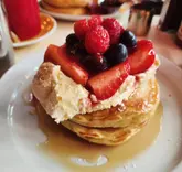 Here are a few options for your alt text, depending on where you want to focus:

**Option 1 (Descriptive & SEO focused):**
Stack of fluffy OG pancakes with vanilla cream, fresh berries, and maple syrup at The Breakfast Club, Liverpool Street.

**Option 2 (Action/Experience focused):**
Enjoying a delicious stack of pancakes with cream and berries for brunch at The Breakfast Club in London.

**Option 3 (Concise):**
Pancakes topped with vanilla cream, fresh berries, and maple syrup at The Breakfast Club, Liverpool Street.

**SEO Tip:** I recommend **Option 1** as it includes the specific location (Liverpool Street) and the signature menu item, both of which are high-intent search terms for food bloggers.