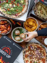 A high-quality, overhead flat-lay photograph showcasing a generous spread of wood-fired pizzas, waffle fries, chicken wings, and arancini from Fired Up Pizza. The image captures a hand reaching for a piece of food, adding a human element to the scene, while the branded pizza boxes clearly identify the venue. The lighting is bright and even, highlighting the textures of the food, making it look fresh and appealing.