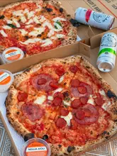 Here are a few options for your SEO alt text, depending on what is actually shown in the photo:

**Option 1 (Focus on the food/drink):**
"People enjoying Rudy’s Pizza, White Claw hard seltzers, and Jubel beer during a sunny park picnic."

**Option 2 (Focus on the vibe):**
"Friends drinking rosé, White Claws, and Jubel beer with Rudy’s pizza during a picnic in a London park."

**Option 3 (Concise):**
"A summer park picnic featuring Rudy’s Pizza, White Claw, and Jubel beer in London."

**SEO Tip:** I included the brand names (Rudy’s, White Claw, Jubel) and the location (London) as these are high-value keywords for search engines and accessibility.