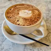 A high-quality, top-down shot of a mocha in a ceramic cup, served on a marble table with a gold and black spoon. The image captures the latte art and the clean, modern aesthetic of the cafe.