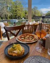 A bright, inviting shot of a lunch setting at a waterside venue, featuring a pizza and an Aperol Spritz. The natural lighting highlights the outdoor dining experience.