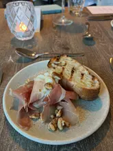 A close-up, high-angle shot of a starter dish featuring prosciutto, burrata, and grilled bread, served on a rustic wooden table in a restaurant setting.