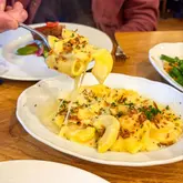 A close-up, high-angle shot of a mac and cheese dish being lifted with a fork, highlighting a cheese pull. The setting is a restaurant table with other plates and a person visible in the background.
