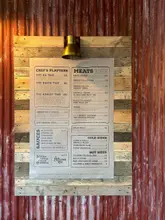 A clear, well-lit photograph of a menu board for a BBQ restaurant, mounted on a rustic corrugated metal and wood wall. The image effectively communicates the restaurant's offerings, pricing, and casual, industrial-style aesthetic.