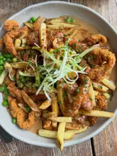 A top-down, well-lit photograph of a plate of loaded fries topped with crispy chicken, peas, a creamy sauce, and fresh spring onions, served on a wooden table.