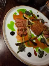 A high-angle shot of a roasted spring lamb dish served with spring onions, black garlic, and a breaded component, presented with artistic sauce drizzles on a white plate.