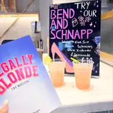 A promotional shot taken at a theatre bar, featuring two plastic cups of a pink cocktail next to a 'Legally Blonde' theatre program, with a handwritten menu board in the background.