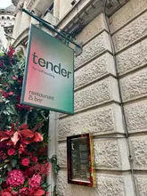 A high-quality, eye-level exterior shot of the signage for 'Tender by Niall Keating', mounted on a stone building facade and framed by a vibrant red floral display.