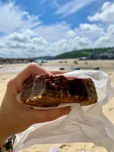 A close-up, handheld shot of a brookie (brownie-cookie hybrid) being held against a blurred beach and sea background.