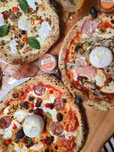 An overhead shot of three pizzas on a wooden table, accompanied by small pots of dips and a cocktail glass, showcasing a casual dining experience at a pizza restaurant.