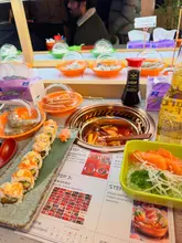 A casual, overhead shot of a conveyor belt hotpot and sushi meal. The image captures the interactive nature of the dining experience, showing the hotpot, sushi, and sashimi spread across the table.