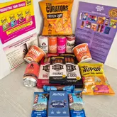 A flat-lay product photography shot showcasing the contents of a 'Hello Yummy Box,' featuring various snacks, drinks, and health-focused food items arranged on a neutral surface.