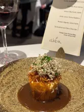 A close-up shot of a fine-dining pastry dish, likely a choux bun, served on a textured plate with a rich sauce. A menu card and a glass of red wine are visible in the background.