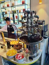 A shot of a chocolate fountain station at a buffet, featuring bowls of fruit, brownies, and marshmallows in the foreground. The image captures the indulgent nature of the dessert section, with the fountain as the focal point.