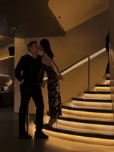 A stylish, atmospheric lifestyle shot of a couple on a staircase at Mr Porter, capturing a romantic date night vibe. The image relies on warm, moody lighting to highlight the venue's interior design and create an intimate, aspirational aesthetic.
