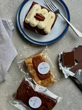 A high-quality, overhead flat-lay photograph showcasing a selection of homemade baked goods, including a slice of carrot cake, a salted caramel brownie, and a Biscoff blondie. The image is styled with a napkin, a fork, and pieces of chocolate on a neutral stone surface, creating an inviting and professional aesthetic.