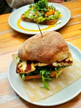 A high-quality, close-up shot of a halloumi sandwich served on branded paper, with an avocado toast dish visible in the background. The lighting is natural and bright, effectively highlighting the textures of the grilled halloumi and toasted bread.