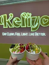 A close-up, eye-level shot of two cups of frozen yogurt held by a creator in front of a branded neon sign. The image focuses on the colourful toppings and the branding of the venue.