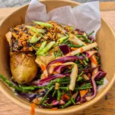 A high-quality, overhead shot of a salmon dish served in a compostable bowl, likely from a street food market. The dish features a piece of cooked salmon topped with crispy onions and spring onions, accompanied by roasted potatoes and a vibrant red cabbage and samphire slaw. The lighting is natural and even, highlighting the textures and colours of the food effectively.