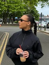 A lifestyle portrait of a person wearing a black Nike/Skims jacket, holding an iced coffee while standing on a street.
