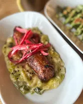 A close-up, top-down shot of a sausage dish served on a bed of leeks with pickled onions, with a side of asparagus visible in the background.