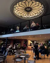 A wide-angle interior shot of a stylish, multi-level hospitality venue featuring a prominent domed ceiling. The space is bustling with customers, showcasing a mix of dining and social areas across a ground floor and a mezzanine level. The atmosphere appears warm and inviting, with a blend of modern and classic architectural elements.