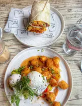A high-quality, overhead flat-lay shot of a brunch spread featuring a plate of smashed avocado on toast topped with poached eggs and crispy halloumi bites, alongside a breakfast burrito. The lighting is natural and bright, effectively highlighting the textures of the food.