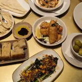 A flat-lay photograph of a tapas spread on a gold-colored table, featuring various dishes including olives, focaccia, hummus, charred broccoli, and crispy sea bass.