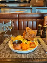 A photograph of a Sunday roast dinner served at a restaurant, featuring a large Yorkshire pudding, roast chicken, and various sides, presented on a table with wine and water glasses.