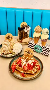 Here are a few options for your SEO alt text, depending on what is most prominent in your photo:

**Option 1 (Focus on the spread):**
A delicious selection of ice cream sundaes and waffles at Llanfaes Dairy in Cardiff Bay.

**Option 2 (Focus on the shop/brand):**
Display of ice cream flavours and indulgent dessert waffles at Llanfaes Dairy, Mermaid Quay.

**Option 3 (Detailed for accessibility):**
Oreo fudge sundae and indulgent waffles served at Llanfaes Dairy ice cream shop in Cardiff Bay.

**Best practice tip:** If your image features a specific person or a clear view of the shop front, replace the first few words with those details to make the description even more accurate!