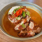 A high-quality, overhead shot of a Thai fried chicken dish served with rice and a generous portion of satay sauce, garnished with fresh coriander and sliced red chili.