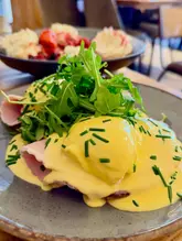 A close-up, eye-level shot of an Eggs Benedict dish featuring poached eggs, hollandaise sauce, and arugula, with a blurred plate of pancakes visible in the background, typical of a cafe brunch setting.