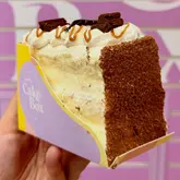 A close-up, eye-level shot of a slice of cake held in a hand, showcasing the texture and layers against a branded purple background.