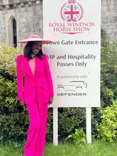 A high-quality lifestyle portrait of a woman in a vibrant pink suit and matching hat, posing next to a Royal Windsor Horse Show sign. The image is bright, clear, and effectively captures the aesthetic of the British social season.