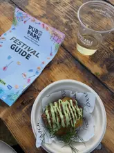 A top-down, natural light shot of a food dish and a drink on a wooden table at a festival, with a festival guide brochure visible.