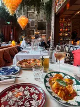 A high-quality, inviting shot of a table at La Bellezza, featuring a pizza and a pasta dish, set against the restaurant's signature maximalist, ornate interior. The warm lighting and well-composed angle create an appealing atmosphere that highlights the dining experience.